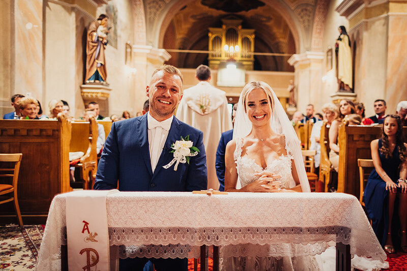 nevesta v závoji pri oltári, ženích v modrom obleku, pár pri kostolnom oltári, svadba v kostole, zlatý organ v pozadí, hostia v laviciach, prirodzené svetlo v chráme. Pálenica Jelšovce.