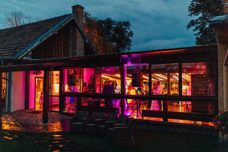 večerná párty v bare, neonové ružové svetlá, hostia tancujú vnútri, modrá obloha pri zotmení, sklenené okná s podsvietením, teplá atmosféra večera. Pálenica Jelšovce.
