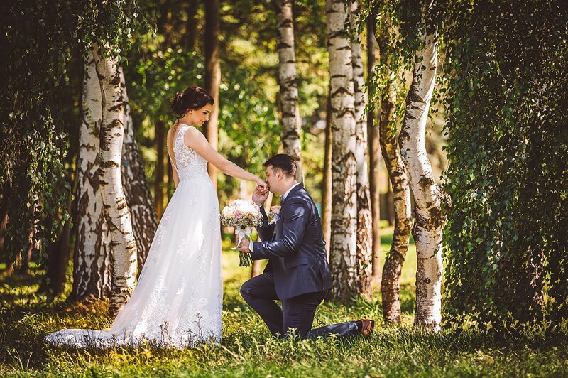 ženích kľačí a bozkáva neveste ruku medzi brezami, vlečka šiat na tráve. Kaštieľ Orlové, Považská Bystrica.