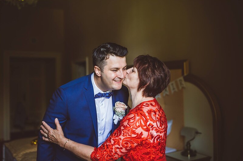 mama v červených šatách bozkáva ženícha na líce, izba v teplom svetle. Kaštieľ Orlové, Považská Bystrica.