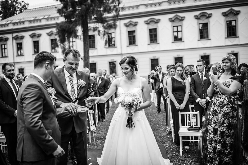 čiernobiely záber, otec odovzdáva nevestu ženíchovi, hostia stoja pozdĺž uličky, fasáda kaštieľa v pozadí. Kaštieľ Orlové, Považská Bystrica.