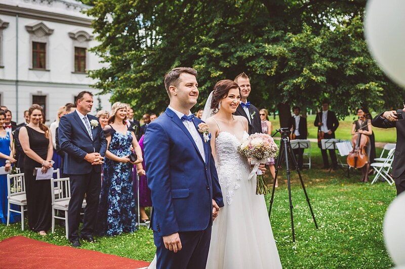 novomanželia stoja na červenom koberci počas obradu, sláčikové kvarteto a hostia v pozadí. Kaštieľ Orlové, Považská Bystrica.