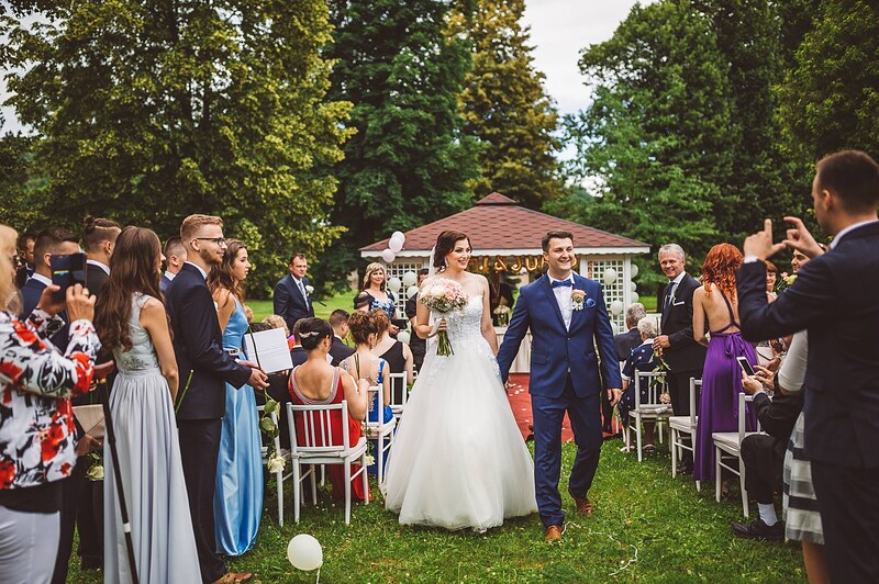 novomanželia odchádzajú uličkou medzi hosťami, altánok a stromy v pozadí, balóny pri stoličkách. Kaštieľ Orlové, Považská Bystrica.