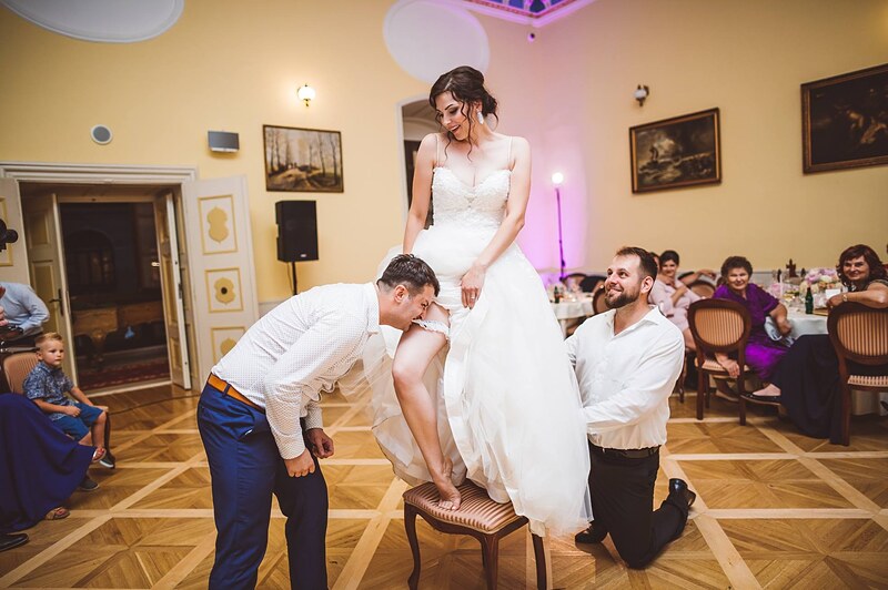ženích sníma neveste podväzok, nevesta stojí na stoličke, svedok kľačí vedľa, hostia sa prizerajú. Kaštieľ Orlové, Považská Bystrica.