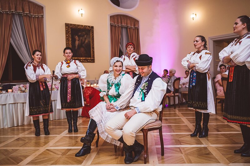 čepčenie, novomanželia sedia na stoličke uprostred folklórneho súboru v krojoch, vyšívané zástery a čižmy. Kaštieľ Orlové, Považská Bystrica.