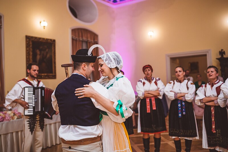 nevesta v čepci a kroji tancuje so ženíchom v klobúku, harmonikár a súbor okolo nich. Kaštieľ Orlové, Považská Bystrica.