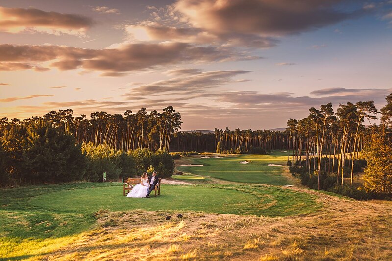 široký záber na golfové ihrisko pri západe slnka, novomanželia ako drobné postavy na lavičke. Penati Golf Resort, Senica.
