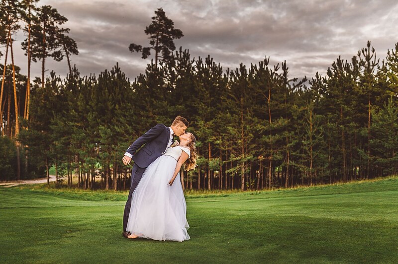 ženích nakláňa nevestu do bozku uprostred ihriska, borovicový les a večerná obloha za nimi. Penati Golf Resort, Senica.