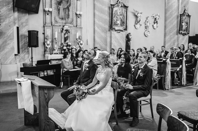 čiernobiely záber z obradu, novomanželia sedia pred oltárom, hostia v laviciach, maľby a sochy na stenách kostola. Fatima, Trenčín.