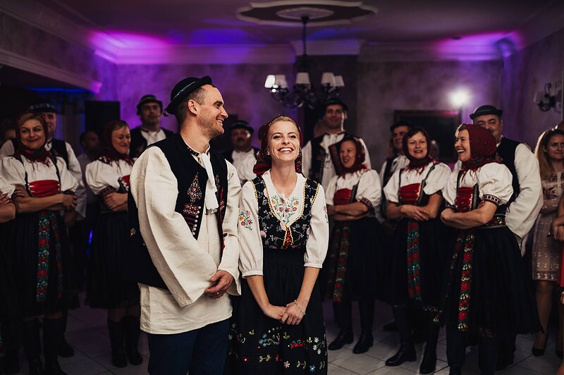 pár v krojoch sa smeje pred folklórnym súborom, vyšívaná zástera a červený pás, hostia v kruhu. Fatima, Trenčín.