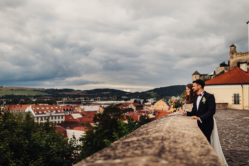 nevesta s kvetmi, ženích v obleku, pár na kamennej stene, výhľad na mesto, červené strechy, oblačné nebo, romantická chvíľa. Fatima, Trenčín.