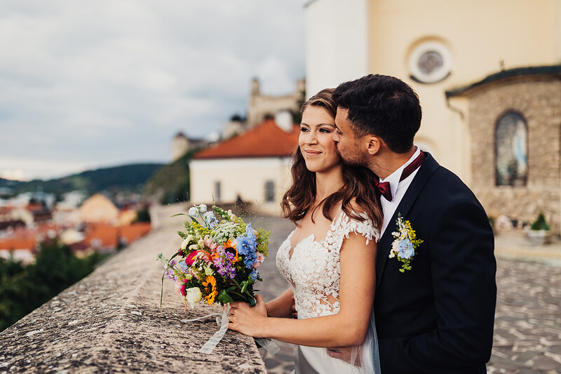 nevesta v čipke, ženích v obleku, pár s kvetmi, historické mesto v pozadí, kamenná terasa, denné svetlo, romantická atmosféra. Fatima, Trenčín.