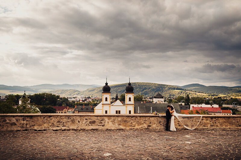 nevesta v bielych šatách, ženích v čiernom obleku, pár na kamennom múre, barokový kostol s kupolami, hornatá krajina v pozadí, oblačné nebo nad scenériou, romantický pohľad do údolia. Fatima, Trenčín.