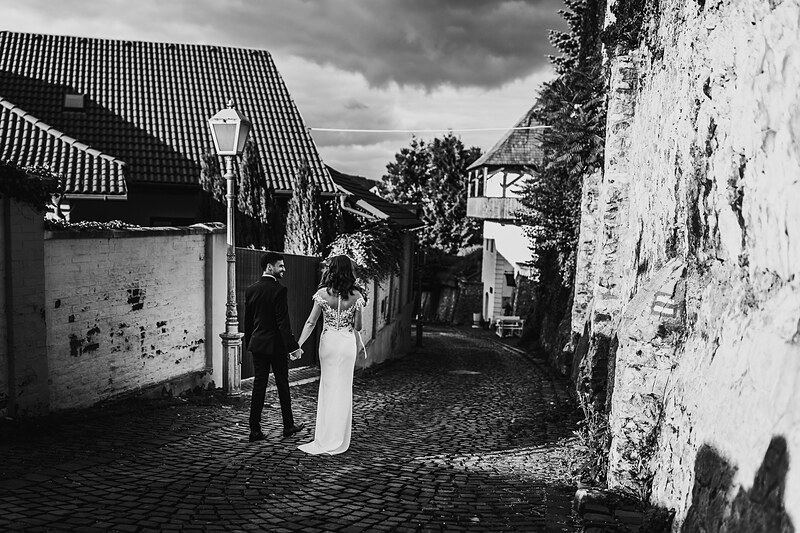 nevesta v bielych šatách, ženích v čiernom obleku, pár držiaci sa za ruky, dlhá ulica s kamennou dlažbou, staré mestské domy, oblačné nebo, vintage architektúra, večerné svetlo, romantická chvíľa. Fatima, Trenčín.
