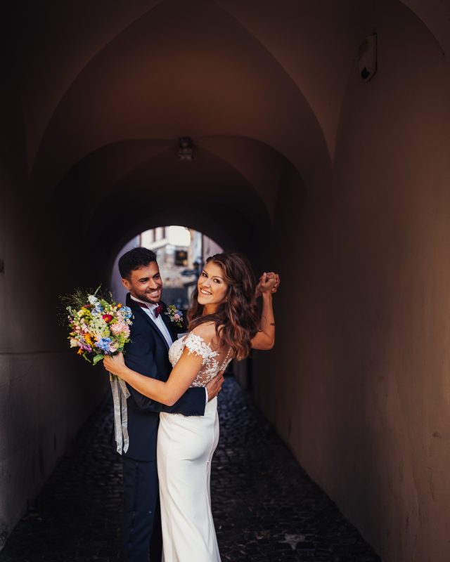 nevesta s kvetmi v tuneli, ženích v tmavom obleku, pár v kamennom oblúku, svetlo z otvoru vzadu, svadobný portrét v tme, úsmev v klenutej chodbe. Fatima, Trenčín.