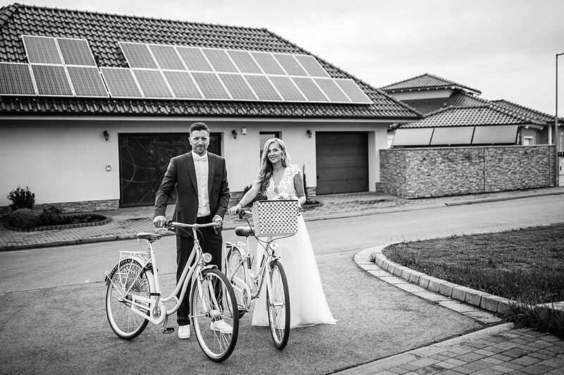 čiernobiely záber, novomanželia s dvoma retro bicyklami na ulici, rodinné domy v pozadí. Gazdovský dvor, Trenčianska Turná.