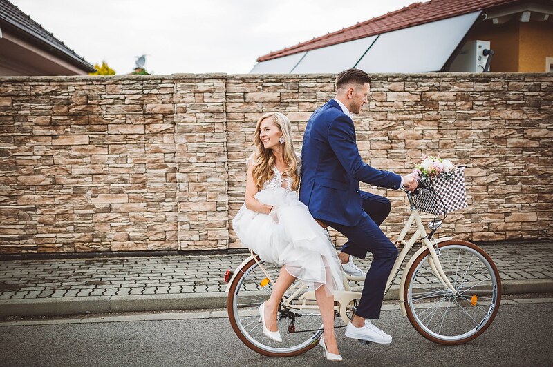 ženích vezie nevestu na ráme bicykla popri kamennom plote, kvety v košíku na riadidlách. Gazdovský dvor, Trenčianska Turná.