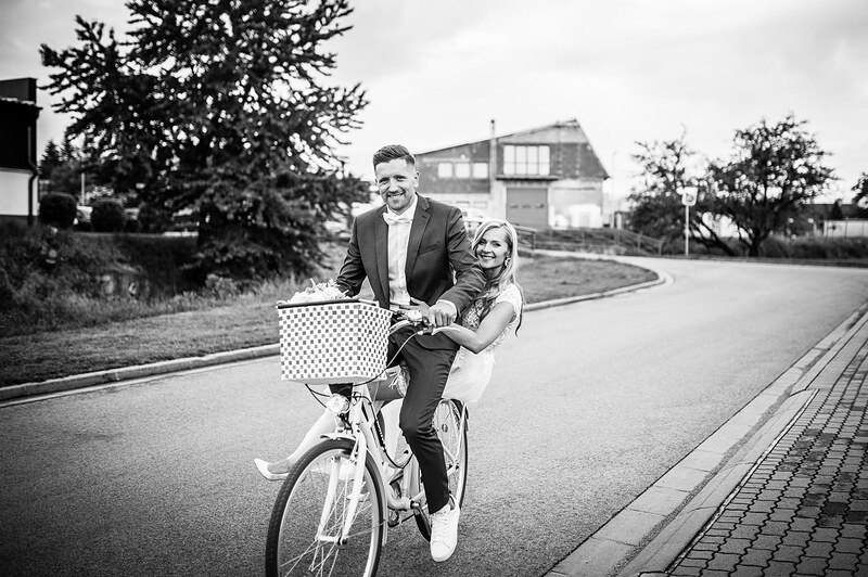 čiernobiely záber, novomanželia sa smejú na jednom bicykli, dedinská cesta a stromy. Gazdovský dvor, Trenčianska Turná.