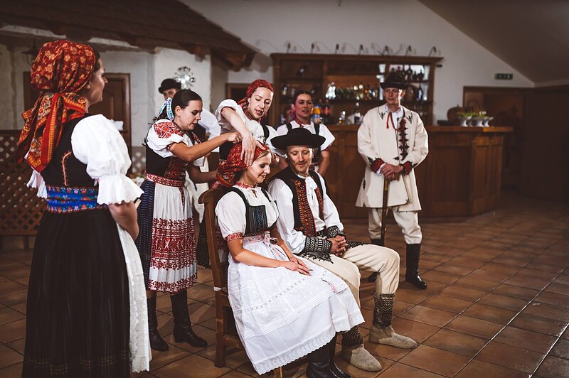 folklórny súbor v krojoch okolo sediacej nevesty, vyšívané zástery a čižmy, biela klenba sály. Gazdovský dvor, Trenčianska Turná.