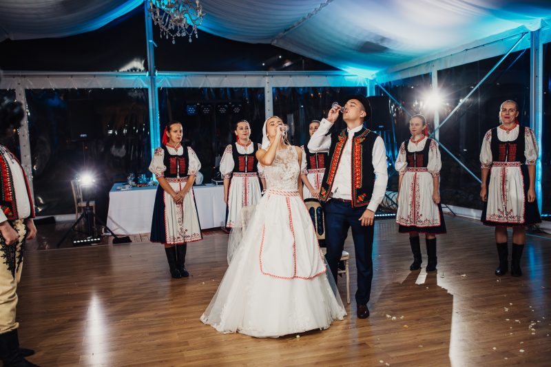 nevesta spieva do mikrofónu počas čepčenia, ženích si pripíja, folklórny súbor v krojoch okolo. Vila Sokolka, Trenčín.