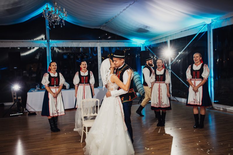 novomanželia v krojoch po čepčení, nevesta v bielom čepci a ženích v klobúku, tanečníci sa smejú. Vila Sokolka, Trenčín.