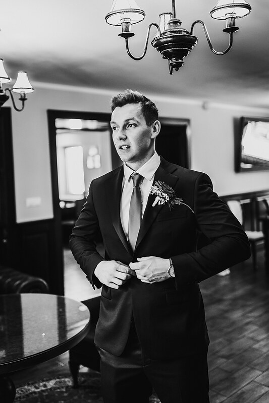 ženích upravujúci kravatu, čiernobiely portrét, muž v obleku s kvetinou, interiér s lustrom, formálne svadobné oblečenie, vážny pohľad ženícha. Vila Sokolka, Trenčín.