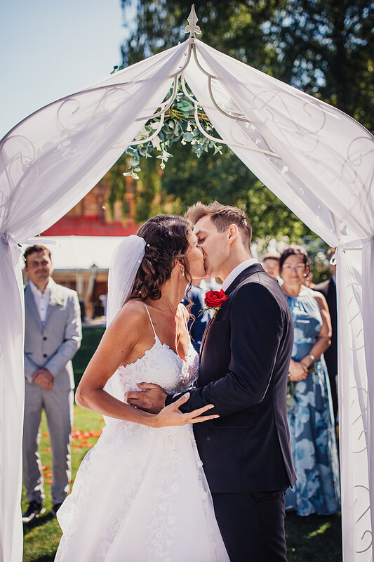 nevesta a ženích sa bozkávajú, biely svadobný stan so zeleňou, svadobný obrad vonku, slnečný deň v záhrade, hostia sledujú prvý bozk, nevesta v čipkovaných šatách. Vila Sokolka, Trenčín.
