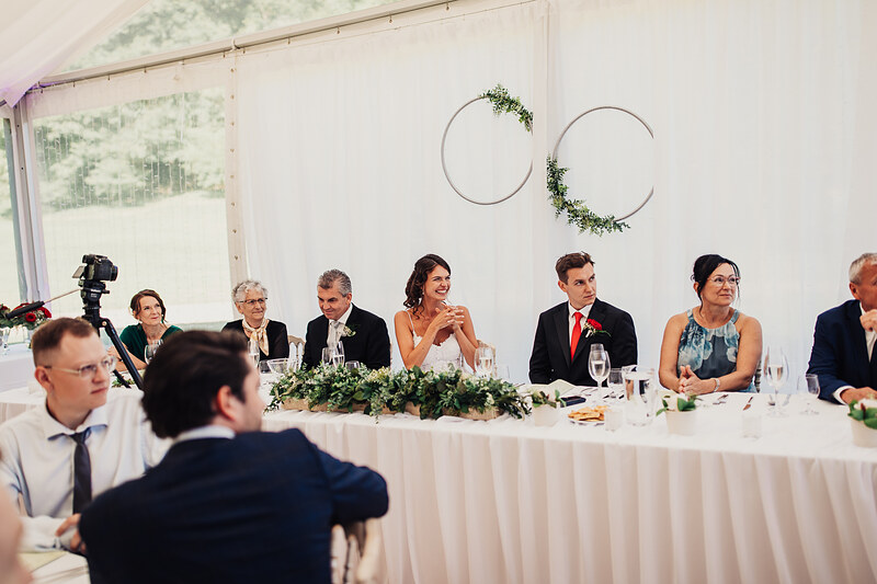 svadobný stôl s hostami, nevesta v oranžových šatách, ženích v červenej kravate, dekorácia so zelenými kruhmi, svetlý stan s oknami, svadobný príhovor, hostia sedia pri stole. Vila Sokolka, Trenčín.
