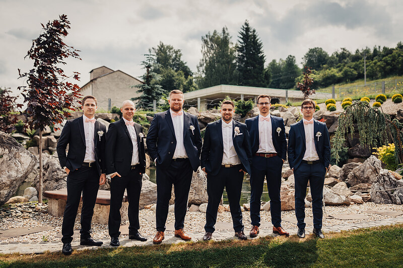 šiesti muži v oblekoch, ženích s kvetinou v klopke, muži stojaci v rade, kamenná záhrada s jazerom, moderný pavilón v pozadí, oblačné nebo nad krajinou. Village Resort Hanuliak.