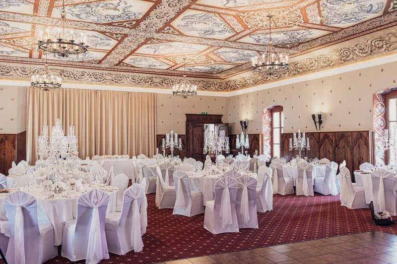 svadobná sála s krištáľovými lustrami, biele stoličky s mašľami, okrúhle stoly s kvetmi, elegantný strop s ornamentami, svadobný banket v príprave, kvetinové dekorácie na stoloch. Zámok Vígľaš.
