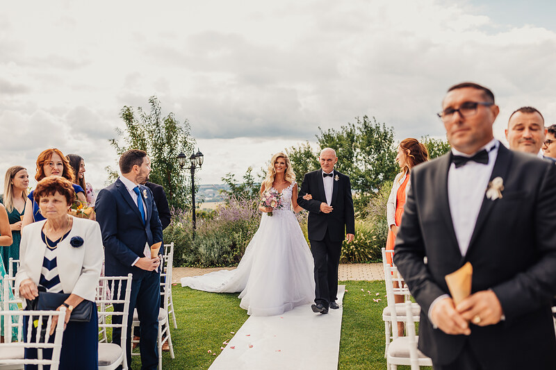 nevesta v bielych šatách, ženích v smokingu, vonkajší obrad na trávniku, hostia v radoch, krajina v pozadí, oblačné nebo, slnečný deň. Hotel Zlatý Kľúčik, Nitra.