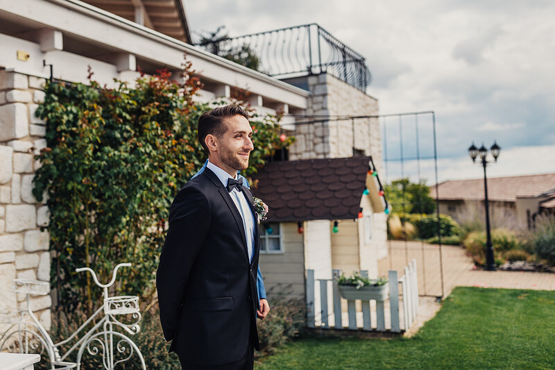 ženích v obleku s motýlikom, muž v čiernom smokingu, svadobný portrét vonku, terasa s kvetinami, pohľad na more v pozadí, slnečný deň s oblakmi. Hotel Zlatý Kľúčik, Nitra.