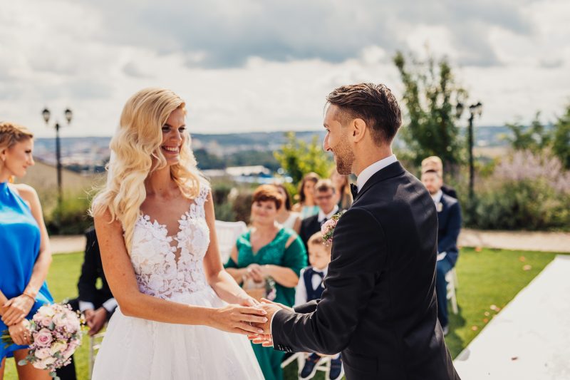 nevesta v bielych šatách, ženích v čiernom obleku, výmena prsteňov, svadobný obrad vonku, krajina s výhľadom, slnečný deň s oblakmi, hostia v pozadí. Hotel Zlatý Kľúčik, Nitra.
