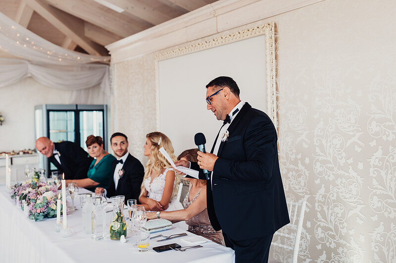 muž s mikrofónom pri stole, svadobní hostia sedia, biele dekorácie na stole, elegantná svadobná sála, príhovor pri večeri, kvetinové aranžmány na stole. Hotel Zlatý Kľúčik, Nitra.