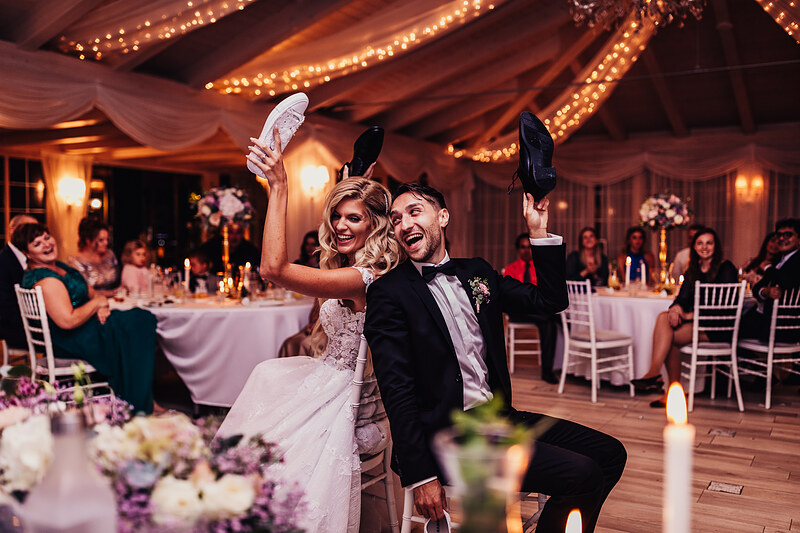 nevesta a ženích s topánkami, svadobný tanec v sále, zlaté osvetlenie a sviečky, hostia pri stoloch, pár so zdvihnutými topánkami, večerná svadobná hostina. Hotel Zlatý Kľúčik, Nitra.