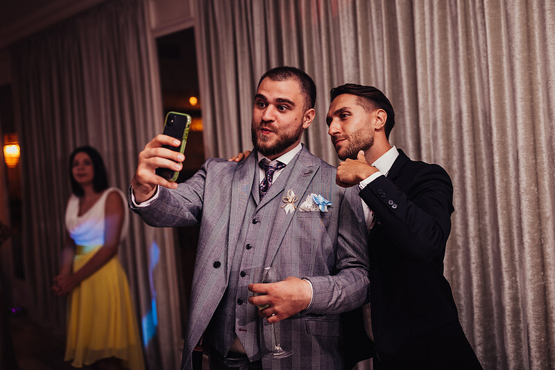 muži v oblekoch si robia selfie, svadobní hostia tancujú, elegantná sála so závesmi, teplé svetlo večera, muž v sivom obleku, formálna oslava v interiéri. Hotel Zlatý Kľúčik, Nitra.