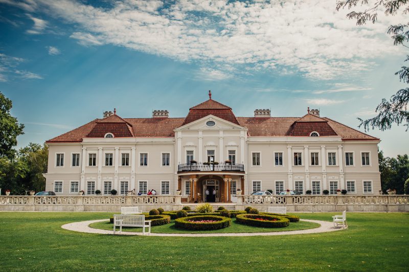 klasický zámok v slnku, biela fasáda s červenou strechou, formálna záhrada s trávnikom, pár pred vchodom, jasný deň s oblakmi, elegantné prostredie na svadbu. Art Hotel Kaštieľ Tomášov.