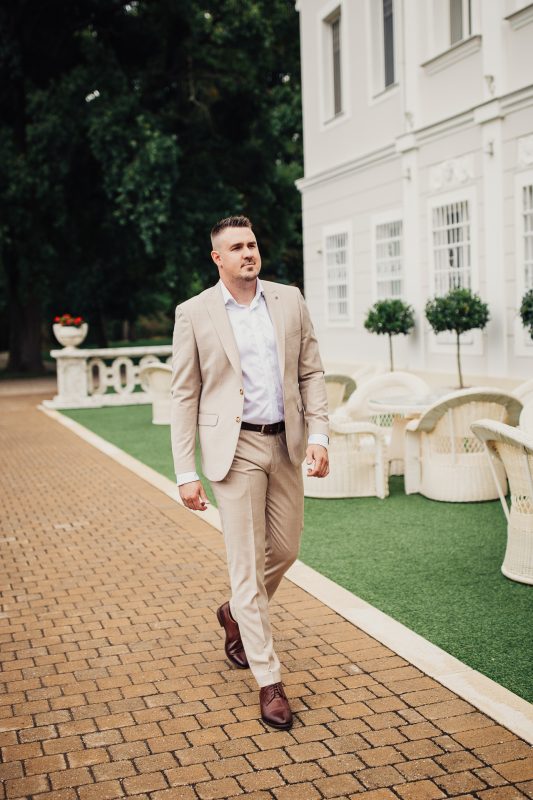 ženích v béžovom obleku, muž kráčajúci po tehlových dlaždicách, biela budova s kolonádou, formálne svadobné oblečenie, denné svetlo v záhrade, elegantný muž v kravate. Art Hotel Kaštieľ Tomášov.