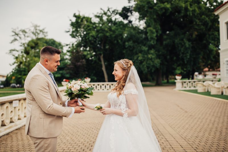 nevesta so závojom, ženích v béžovom obleku, svadobná kytica, dlhá biela róba, tehlová terasa, zelené stromy v pozadí, pár si podáva ruky, dážď padá, denné svetlo, usmiati mladomanželia. Art Hotel Kaštieľ Tomášov.