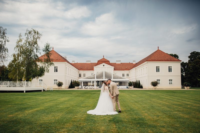 nevesta a ženích sa bozkávajú, západ slnka nad trávnikom, svadobný pár pred palácom, červené strechy a biele steny, denné svetlo a oblaky, nevesta v dlhom závoji. Art Hotel Kaštieľ Tomášov.