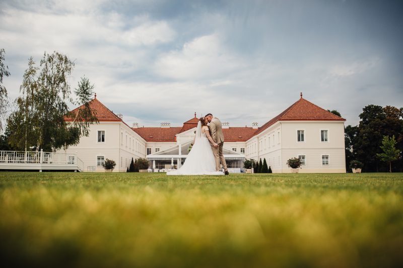 ženích a nevesta na trávniku, biele svadobné šaty, klasický zámok v pozadí, poludňajšie svetlo, romantický bozk, pár na zelenom poli. Art Hotel Kaštieľ Tomášov.