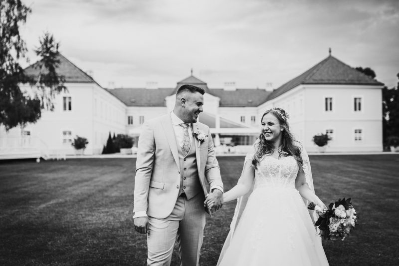 nevesta a ženích držia sa za ruky, pár kráča po trávniku, svadobný kvet v ruke, klasický zámok v pozadí, slnečný deň s oblakmi. Art Hotel Kaštieľ Tomášov.