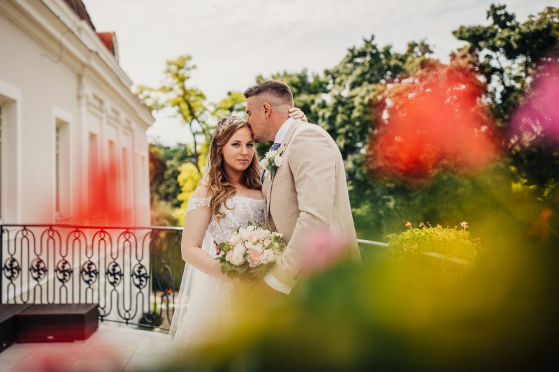 nevesta s kvetmi, ženích bozkáva nevestu, terasa so zábradlím, červené kvety v pozadí, denné svetlo, romantická chvíľa. Art Hotel Kaštieľ Tomášov.