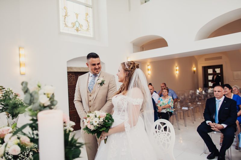 nevesta v bielych šatách, ženích v béžovom obleku, slávnostný obrad v interiéri, sviečky a zlaté svetlá, rodina sedí v pozadí, mladý pár si vymieňa pohľady. Art Hotel Kaštieľ Tomášov.