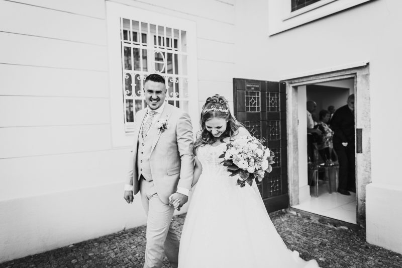 nevesta s kvetmi, ženích v svetlom obleku, pár držiaci sa za ruky, biela architektúra, svadba pred domom. Art Hotel Kaštieľ Tomášov.