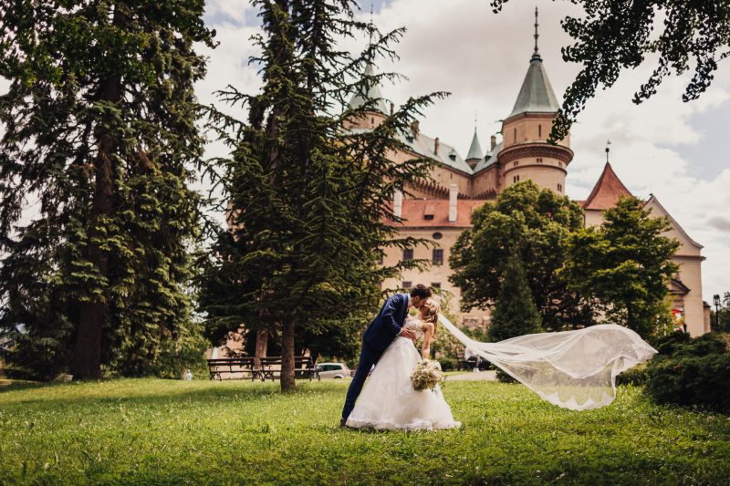 ženích drží nevestu, nevesta v bielych šatách, svadobný tanec na trávniku, hrad v pozadí, dlhý závoj vo vetre, párik pod stromami. Meridiana Resort, Bojnice.