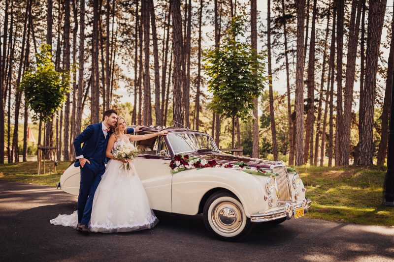 nevesta v bielych šatách, ženích v modrom obleku, vintage auto s kvetmi, pár pri starom aute, lesná alej so stromami, slnečný deň v prírode, mladomanželia pri kabriolete. Meridiana Resort, Bojnice.