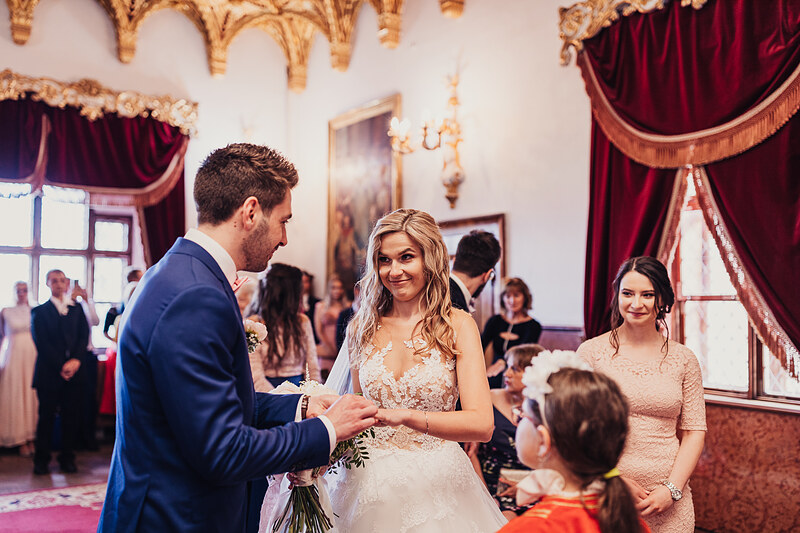 nevesta v čipke, ženích v modrom obleku, výmena prsteňov, slávnostná sála so zlatými dekoráciami, červené závesy, hostia v pozadí. Meridiana Resort, Bojnice.