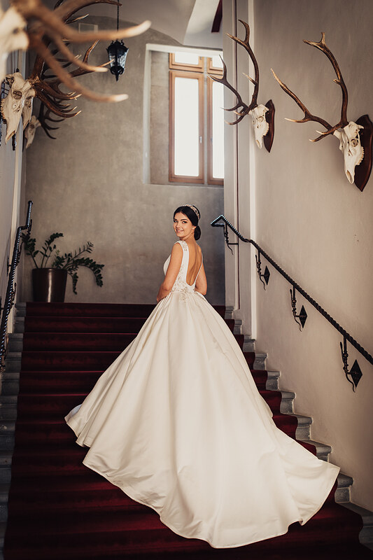 nevesta v bielych šatách, dlhá vlečka na schodoch, lovecké trofeje na stene, prírodné svetlo z okna, elegantný interiér so schodiskom, nevesta pohľadom cez plece. Zámok Vígľaš.