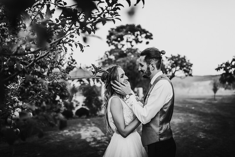 nevesta v bielych šatách, ženích v sivej veste, pár medzi ovocnými stromami, čiernobiely portrét, slnečný deň na vidieku, romantický moment v sade. Karolinenhof Farm, Hainburg.